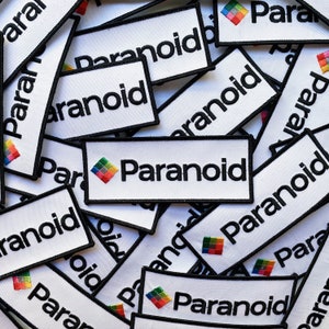 May include: A close-up of several white fabric patches with black text that reads "Paranoid" and a small rainbow logo. The patches are stacked on top of each other.