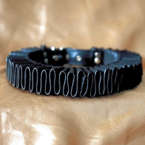 Könnte beinhalten: Ein schwarzes, kreisförmiges Halsband mit einem gefalteten, gerüschten Außenrand und einem glatten Innenband. Das Halsband verfügt über sichtbare silberfarbene Metallbeschläge, darunter eine Schnalle und Einstelllöcher am Innenumfang.