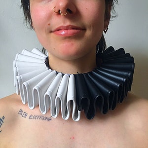 Könnte beinhalten: Ein schwarz-weißer Rüschenkragen aus Kunstleder. Der Kragen wird von einer Person mit einem Nasenring getragen.