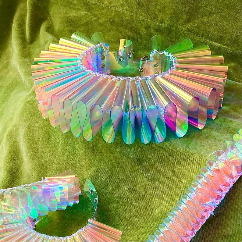 May include: A iridescent, plastic, ruffled collar with a silver clasp. The collar is made of many overlapping, semi-circular pieces of plastic that create a ruffled effect. The collar is a popular fashion accessory for costume and cosplay.
