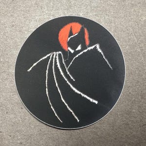 May include: A circular sticker featuring a stylised black silhouette of a figure with a pointed head and a flowing cape. The figure is set against a solid black background with a bright orange circular accent behind the head and white outlines.