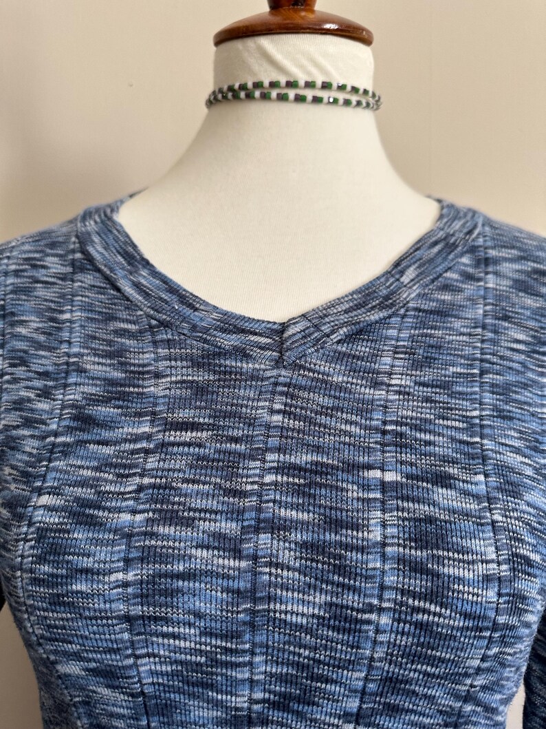 May include: A blue and white ribbed knit top with a V-neck. The fabric has a textured appearance. A beaded choker necklace with green and white beads is around the mannequin's neck.