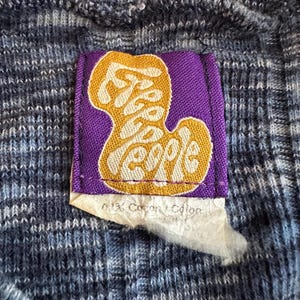 May include: Close-up of a blue and white knit fabric with a purple tag. The tag features the words "Free People" in a gold, stylized font. A small piece of paper with text is attached to the tag.