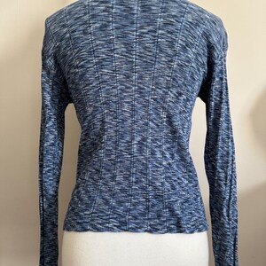 May include: A blue long-sleeve sweater with a ribbed texture. The sweater has a scoop neckline and a fitted silhouette. The fabric has a subtle, heathered appearance, with shades of blue and white. The sleeves are long and fitted.