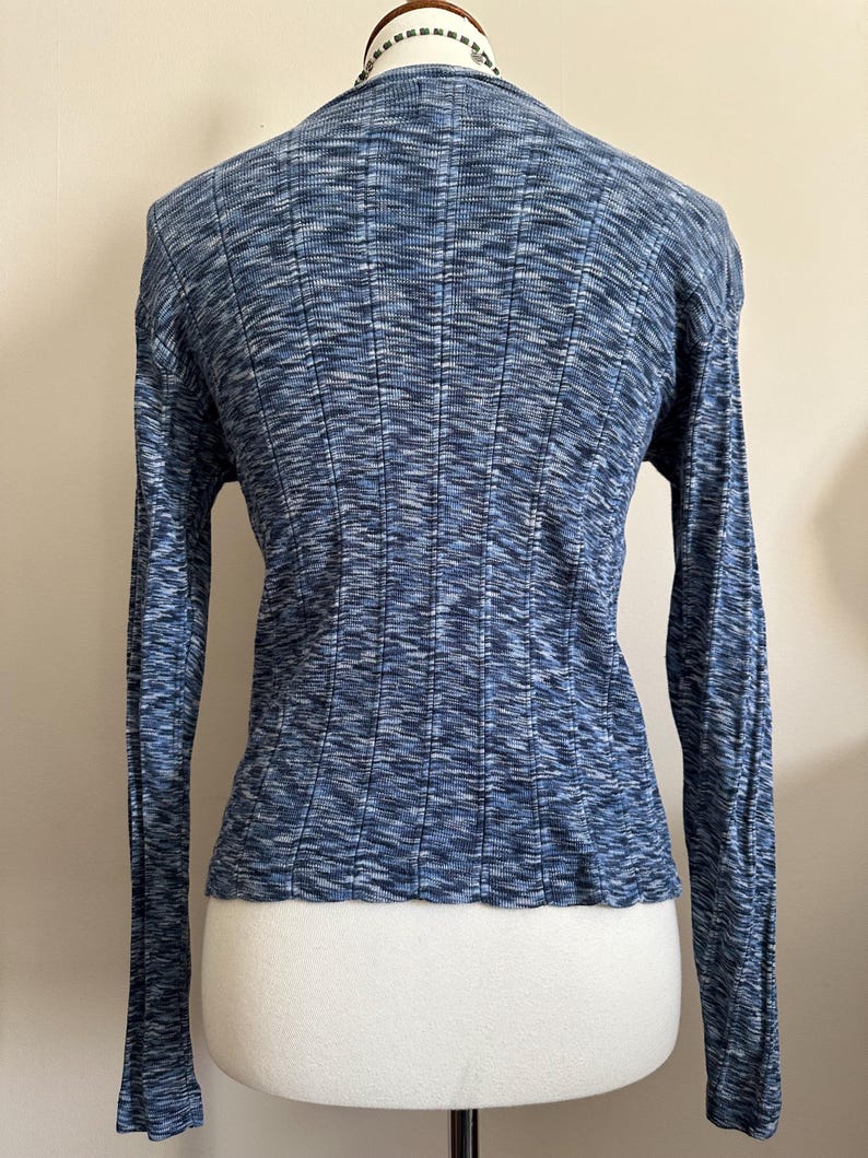 May include: A blue long-sleeve sweater with a ribbed texture. The sweater has a scoop neckline and a fitted silhouette. The fabric has a subtle, heathered appearance, with shades of blue and white. The sleeves are long and fitted.