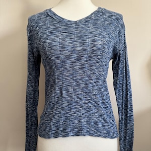 May include: A long-sleeved blue knit shirt with a ribbed texture. The shirt has a V-neck and is displayed on a mannequin. The fabric has a subtle pattern of light and dark blue threads, creating a textured appearance. The sleeves are long and fitted.