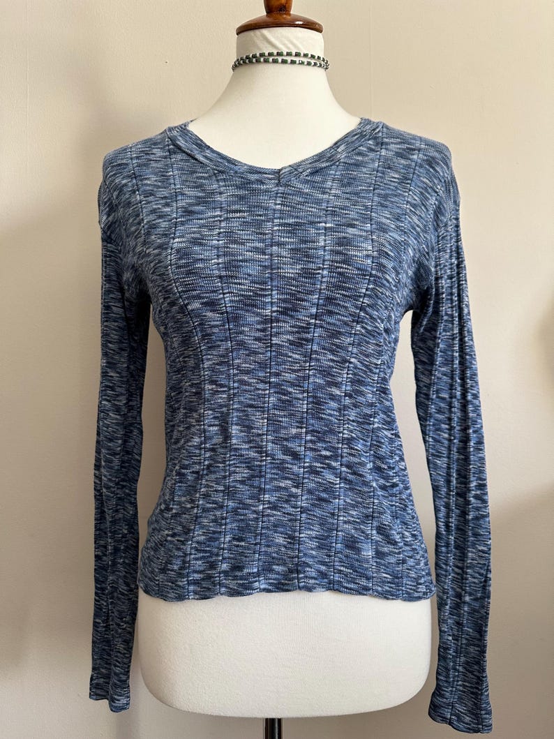 May include: A long-sleeved blue knit shirt with a ribbed texture. The shirt has a V-neck and is displayed on a mannequin. The fabric has a subtle pattern of light and dark blue threads, creating a textured appearance. The sleeves are long and fitted.