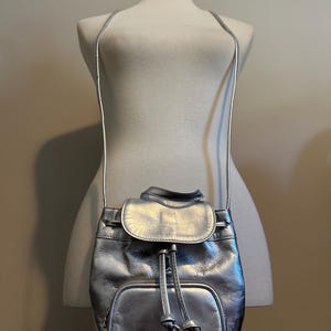 May include: Silver backpack purse with a drawstring closure and a flap. The purse has a small front pocket and thin shoulder straps. The bag is displayed on a mannequin.