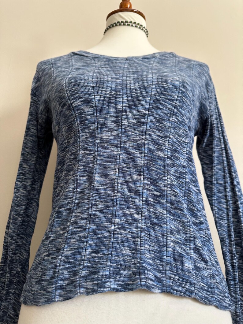 May include: A blue long-sleeve shirt with a ribbed texture. The shirt has a scoop neck and a fitted silhouette. The fabric has a subtle, mottled pattern in shades of blue and white.