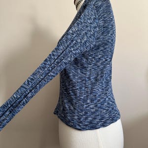 May include: A blue long-sleeve top with a ribbed texture, displayed on a mannequin. The top features a V-neck and a fitted silhouette. The fabric has a mix of blue shades, creating a textured appearance. The sleeves are long and form-fitting.