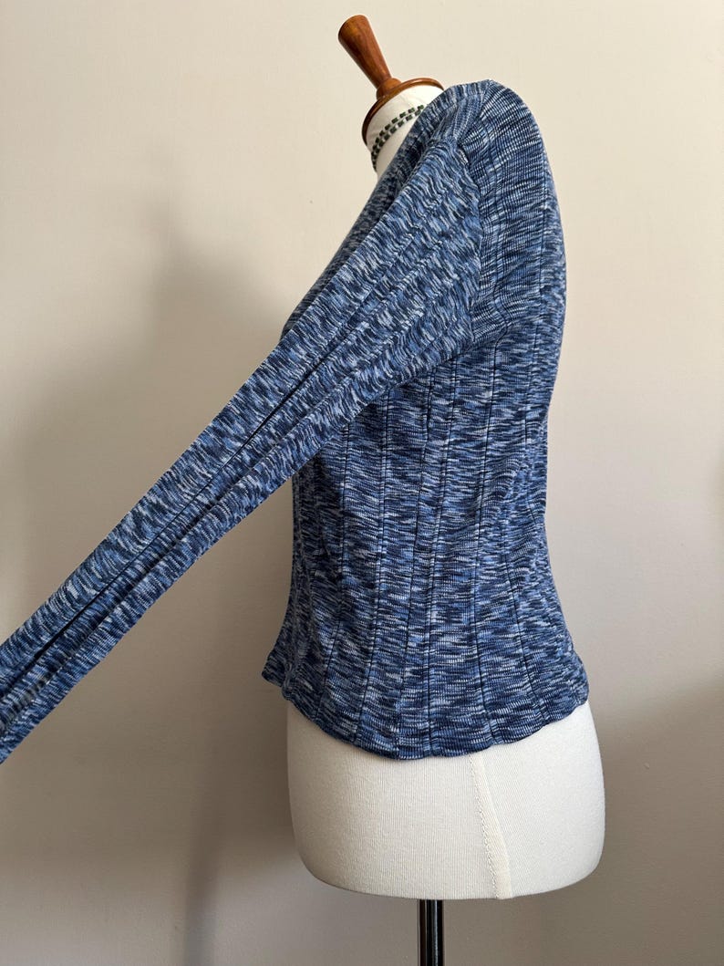 May include: A blue long-sleeve top with a ribbed texture, displayed on a mannequin. The top features a V-neck and a fitted silhouette. The fabric has a mix of blue shades, creating a textured appearance. The sleeves are long and form-fitting.
