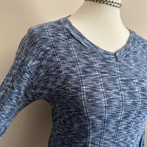 May include: A blue and white marled knit top with a V-neck and ribbed texture. The long-sleeved shirt is displayed on a mannequin. The fabric has a subtle pattern and a soft appearance.
