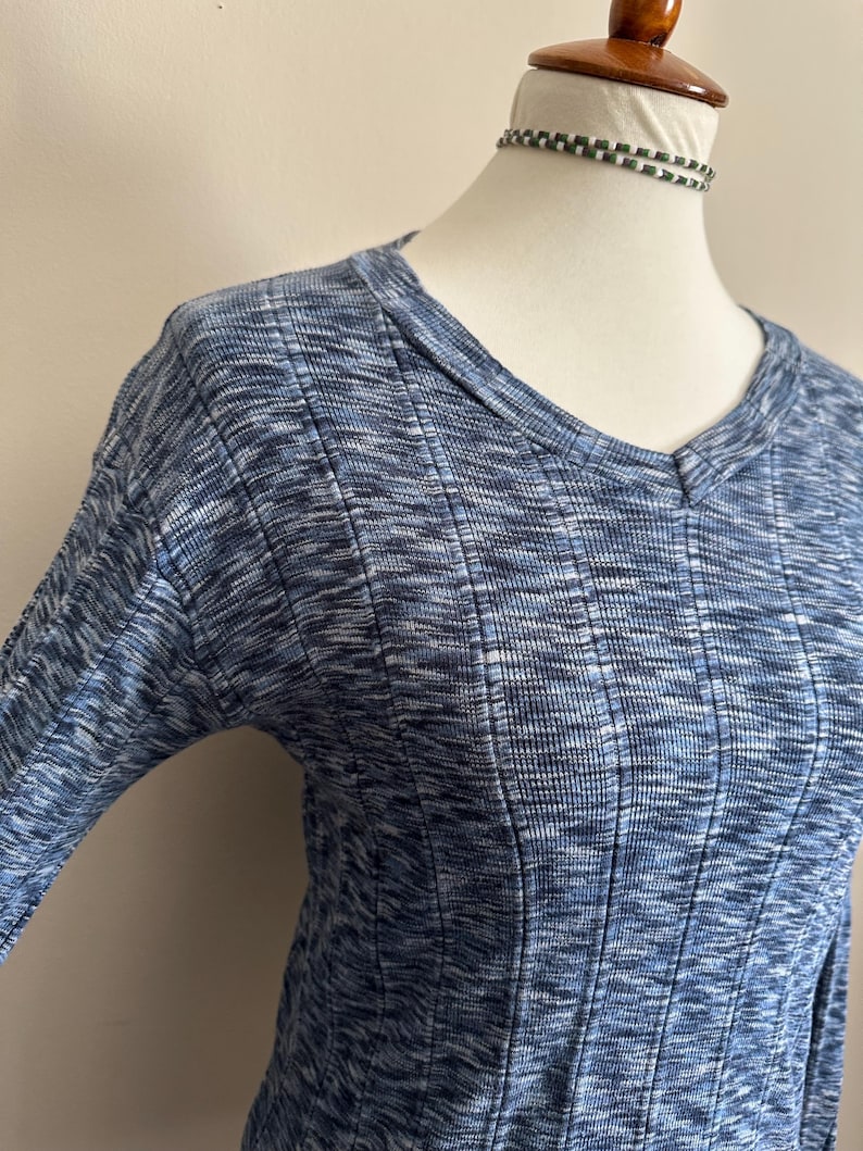 May include: A blue and white marled knit top with a V-neck and ribbed texture. The long-sleeved shirt is displayed on a mannequin. The fabric has a subtle pattern and a soft appearance.