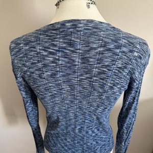 May include: A blue long-sleeve sweater with a ribbed texture. The sweater has a scoop neck and a fitted silhouette. The fabric has a subtle marled effect, with shades of blue and white. A beaded necklace is visible at the neckline.