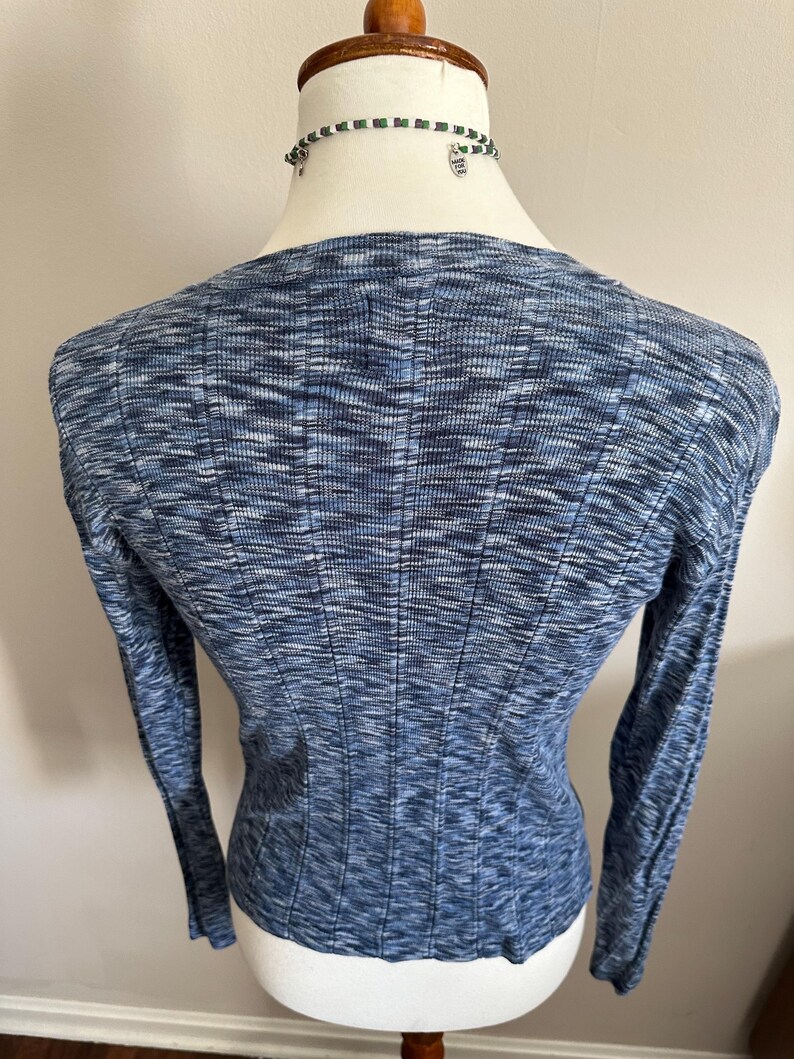 May include: A blue long-sleeve sweater with a ribbed texture. The sweater has a scoop neck and a fitted silhouette. The fabric has a subtle marled effect, with shades of blue and white. A beaded necklace is visible at the neckline.