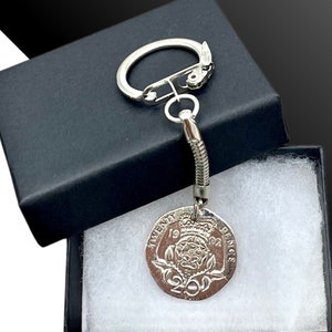 Puede incluir: Un llavero plateado con una moneda británica de dos libras de 2022 adjunta. La moneda presenta un diseño de una corona y una rosa. El llavero está en una caja negra con acolchado blanco.