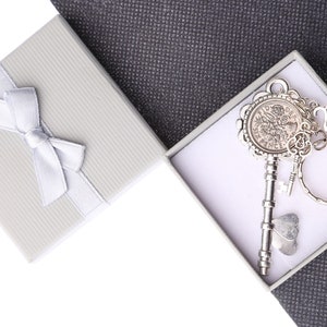 May include: A silver key with a decorative round head and a small charm attached to a silver ring. The key is in a white gift box with a white ribbon.