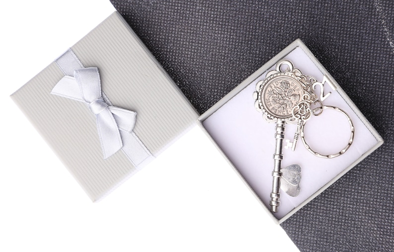May include: A silver key with a decorative round head and a small charm attached to a silver ring. The key is in a white gift box with a white ribbon.