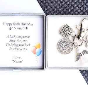 May include: A silver gift box with a card that reads "Happy 80th Birthday" and "A lucky sixpence just for you to bring you luck in all you do". The box contains a silver keychain with a sixpence coin, a birthday cake charm, a heart charm that says "Grandma", and angel wings.