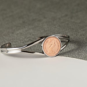 May include: A silver-toned cuff bracelet featuring a copper coin at its center. The coin is set in a circular frame and is the focal point of the open-ended design. The bracelet rests on a white surface with a gray backdrop.