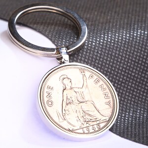 Peut inclure: Porte-clés en argent avec une pièce de monnaie britannique d'un penny de 1940. La pièce représente une femme tenant un trident avec les mots "ONE PENNY" autour du bord.