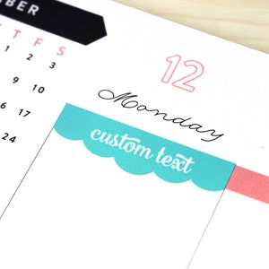 May include: A teal blue calendar sticker with scalloped edges and the text "custom text" in white. The sticker is placed on a calendar page with the number 12 and the word "Monday" written in black.