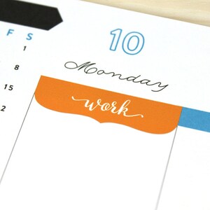 May include: A calendar page with the number 10 circled in blue and the word "Monday" written in black cursive. A bright orange tab with a rounded edge and the word "work" written in white cursive is attached to the page.