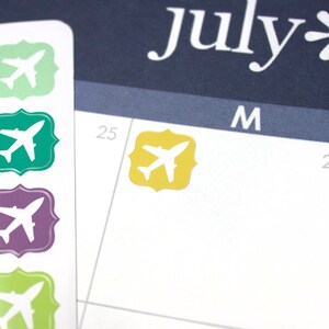 May include: A sheet of colourful airplane stickers on a white background. The stickers are in various colours, including pink, green, yellow, and purple. One sticker is placed on a calendar page with the word "July" visible.