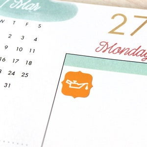 Pode incluir: Uma p&aacute;gina de planejador em close-up com um calend&aacute;rio de mar&ccedil;o. A data "27" e a palavra "Monday" est&atilde;o em ouro e escrita vermelha. Um adesivo laranja com um &iacute;cone branco de lata de &oacute;leo est&aacute; no canto.