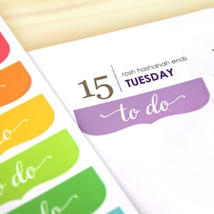 May include: Close-up of colorful planner stickers with the words "to do" in a cursive font. The stickers are in shades of pink, red, orange, yellow, and green. A planner page shows the date "15" and the word "Tuesday" with the text "to do" on a purple sticker.