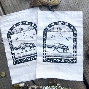 May include: Two white fabric patches with black ink illustrations of a fox walking through a forest scene. The fox is surrounded by birds, trees, and mushrooms. The text "Maya Butterfield" is printed in the bottom right corner of the right patch.