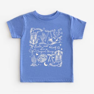 May include: A blue t-shirt with a white graphic design featuring cowboy boots, a horseshoe, a cowboy hat, cacti, and the words "Boots and Bling, it's a Cowgirl thing." The design includes stars, bows, and a crescent moon.
