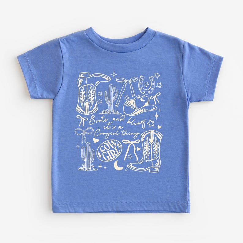 May include: A blue t-shirt with a white graphic design featuring cowboy boots, a horseshoe, a cowboy hat, cacti, and the words "Boots and Bling, it's a Cowgirl thing." The design includes stars, bows, and a crescent moon.