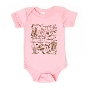 May include: Pale pink baby bodysuit featuring a brown graphic. The design includes cowboy boots, a cowboy hat, a horseshoe, cacti, and the text "Boots and bling it's a Cowgirl thing."