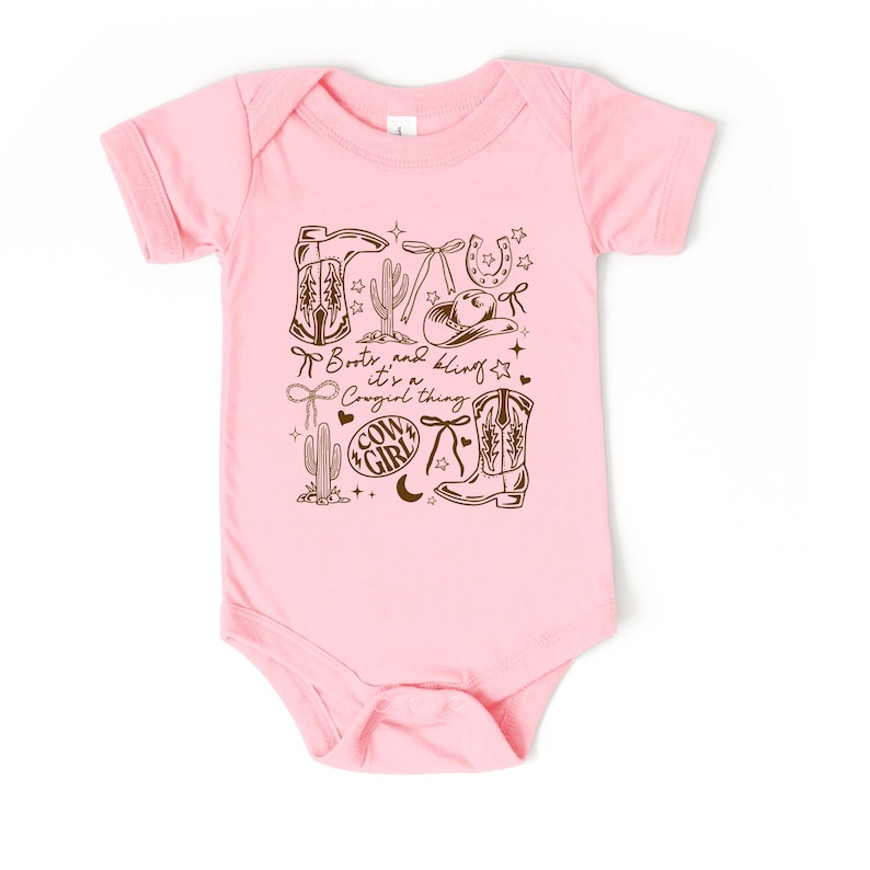May include: Pale pink baby bodysuit featuring a brown graphic. The design includes cowboy boots, a cowboy hat, a horseshoe, cacti, and the text "Boots and bling it's a Cowgirl thing."