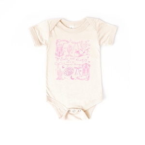 May include: A cream-coloured baby bodysuit featuring a pink cowgirl-themed design. The design includes boots, a horseshoe, a cactus, and the text "Boots and bling, it's a cowgirl thing."