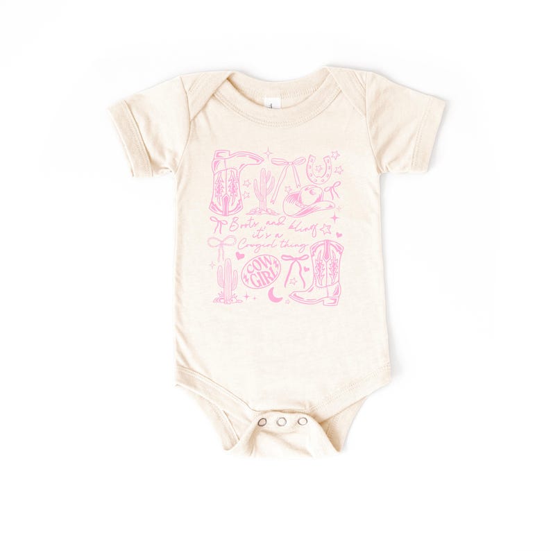 May include: A cream-coloured baby bodysuit featuring a pink cowgirl-themed design. The design includes boots, a horseshoe, a cactus, and the text "Boots and bling, it's a cowgirl thing."