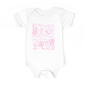 May include: White baby bodysuit featuring a pink cowgirl design. The design includes illustrations of cowboy boots, a hat, a horseshoe, and the text "Boots and bling it's a Cowgirl thing."
