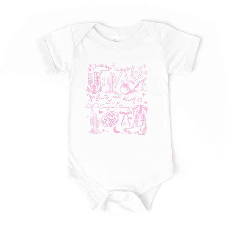 May include: White baby bodysuit featuring a pink cowgirl design. The design includes illustrations of cowboy boots, a hat, a horseshoe, and the text "Boots and bling it's a Cowgirl thing."