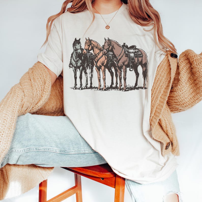 May include: Cream-colored t-shirt featuring a detailed illustration of three horses. The horses are depicted in shades of gray, brown, and tan, with one saddled. The graphic is set against a neutral background, creating a rustic, Western-inspired aesthetic.