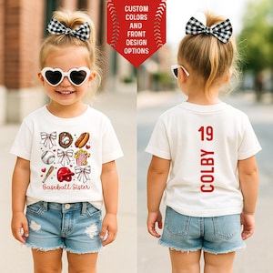 Op de afbeelding: Een kind draagt een wit T-shirt met honkbalthema-afbeeldingen en de tekst "Baseball Sister". De achterkant van het shirt toont de naam "Colby" en het nummer "19". Het kind draagt ook een spijkerbroekshort en een hartvormige zonnebril.