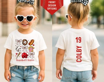 May include: A white short-sleeved t-shirt featuring a front graphic of baseball icons, bows, and the text "Baseball Sister." The back displays the number "19" and the name "COLBY" in red text. The shirt is paired with blue denim shorts.