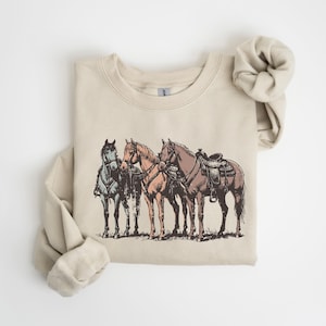 May include: A beige sweatshirt featuring a detailed illustration of three horses. The horses are in shades of blue, tan, and brown, with one saddled. The design has a vintage, sketched aesthetic, evoking a Western or equestrian theme.