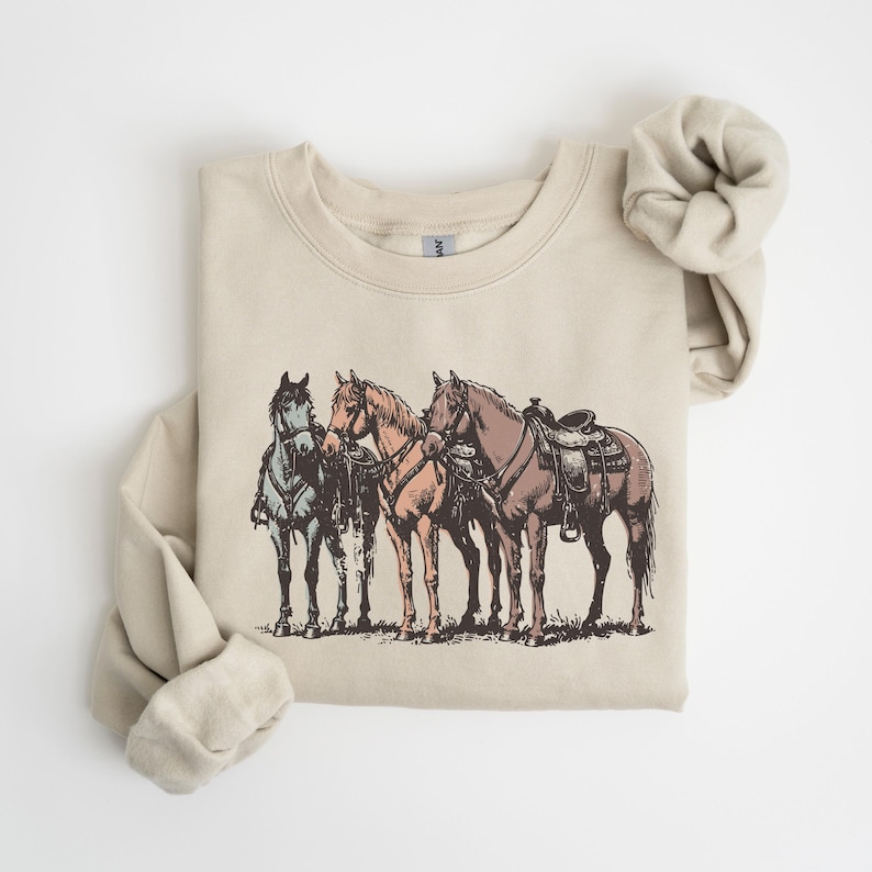 May include: A beige sweatshirt featuring a detailed illustration of three horses. The horses are in shades of blue, tan, and brown, with one saddled. The design has a vintage, sketched aesthetic, evoking a Western or equestrian theme.
