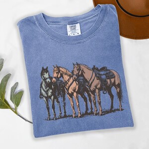 May include: A blue Comfort Colors t-shirt featuring a graphic of three horses with saddles. The horses are in shades of brown and tan, with one horse in green. The shirt has a crew neck and the Comfort Colors label is visible.