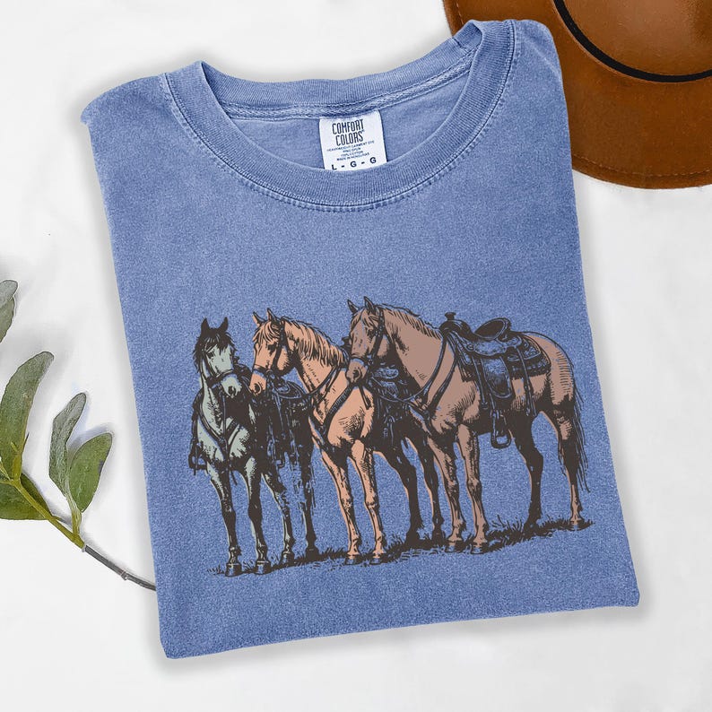 May include: A blue Comfort Colors t-shirt featuring a graphic of three horses with saddles. The horses are in shades of brown and tan, with one horse in green. The shirt has a crew neck and the Comfort Colors label is visible.