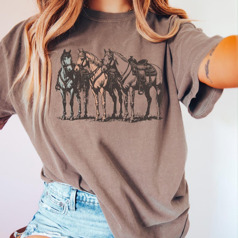 May include: A brown t-shirt featuring a detailed illustration of four horses with saddles and tack. The horses are standing side-by-side on a grassy surface. The shirt is a muted brown color and has a relaxed fit.