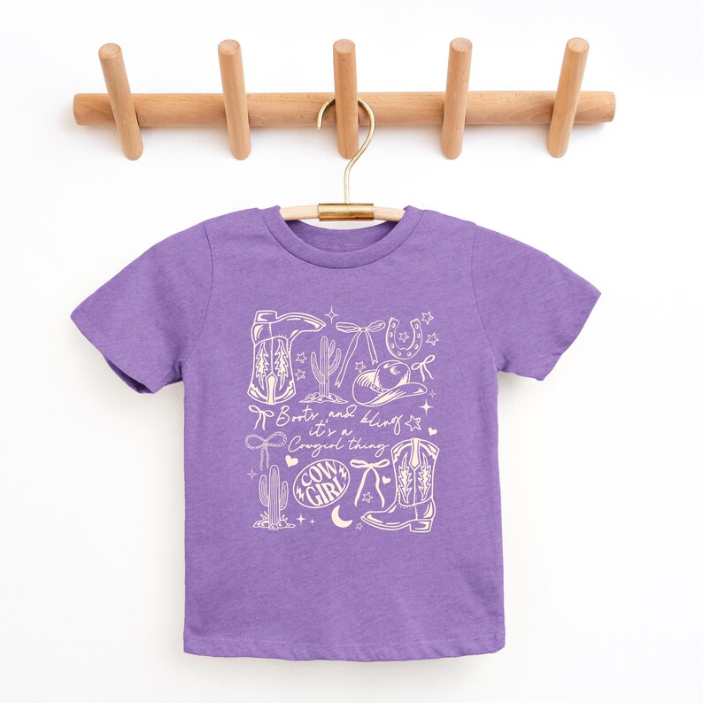 May include: A lilac t-shirt with a cream-coloured graphic design featuring cowboy boots, a cactus, a horseshoe, and the text "Boots and Bling, it's a Cowgirl thing." The shirt is hanging on a wooden hanger.
