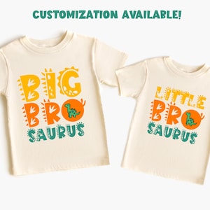 以下が含まれることがあります： 恐竜をテーマにしたグラフィックが特徴の、クリーム色のTシャツ2枚。「BIG BRO SAURUS」はオレンジ、黄色、ティール色。「LITTLE BRO SAURUS」は同様の色。上部のテキストは「カスタマイズ可能！」