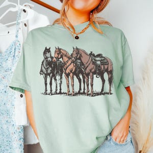 May include: A light green t-shirt featuring a detailed illustration of three horses. The horses are depicted in shades of brown and gray, with one saddled. The design has a vintage, sketched aesthetic, evoking a Western or equestrian theme.